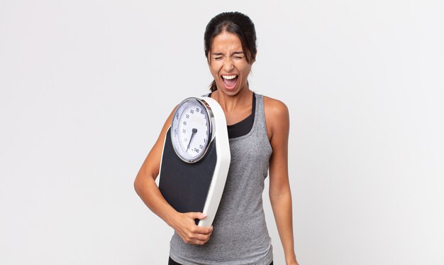 Young hispanic woman shouting aggressively, looking very angry and holding a weight scale. diet concept