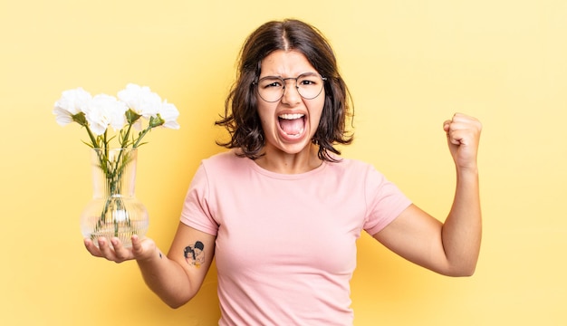 Young hispanic woman shouting aggressively with an angry expression. flowers pot concept