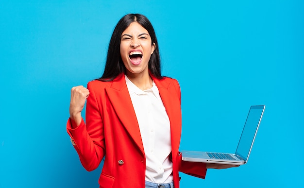 Young hispanic woman shouting aggressively with an angry expression or with fists clenched celebrating success. laptop