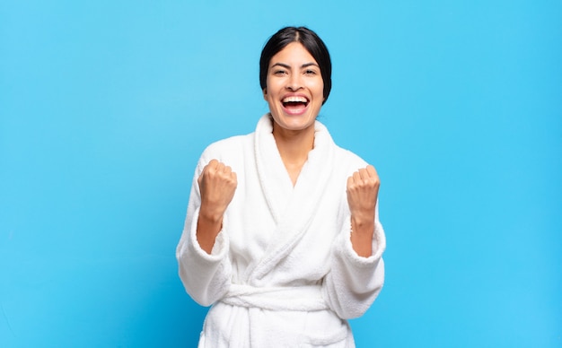 Young hispanic woman shouting triumphantly, laughing and feeling happy and excited while celebrating success. bathrobe concept