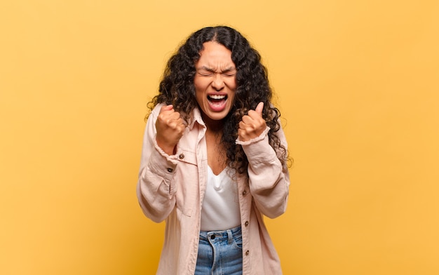 Young hispanic woman shouting triumphantly, laughing and feeling happy and excited while celebrating success