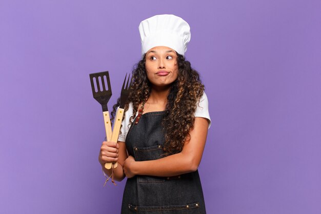 Photo young hispanic woman shrugging, feeling confused and uncertain, doubting with arms crossed and puzzled look. barbecue chef