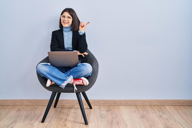 Young hispanic woman sitting on chair using computer laptop with a big smile on face, pointing with hand finger to the side looking at the camera.