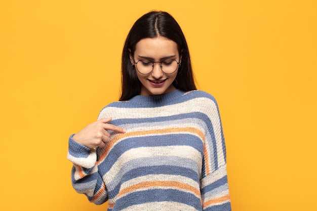 Young hispanic woman smiling cheerfully and casually, looking downwards and pointing to chest