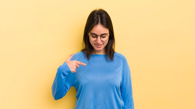 Young hispanic woman smiling cheerfully and casually looking downwards and pointing to chest