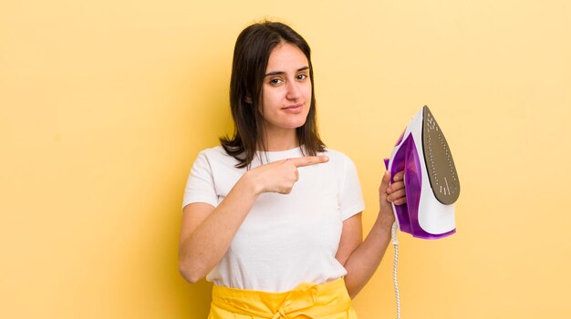 Young hispanic woman smiling cheerfully feeling happy and pointing to the side clothes iron concept