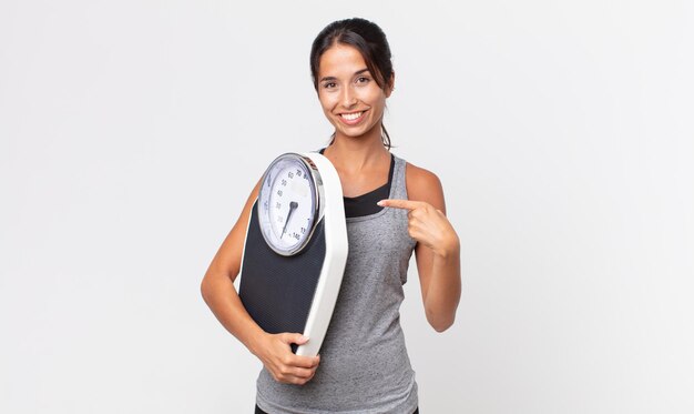 Young hispanic woman smiling cheerfully, feeling happy and pointing to the side and holding a weight scale. diet concept