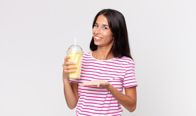 Young hispanic woman smiling cheerfully, feeling happy and showing a concept and holding a vanila smoothy milkshake