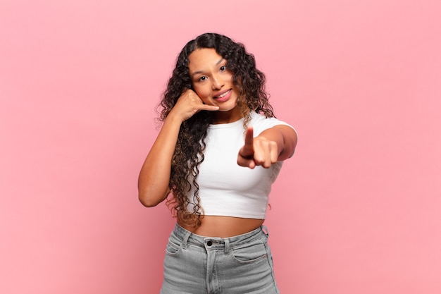 Young hispanic woman smiling cheerfully and pointing to camera while making a call you later gesture, talking on phone