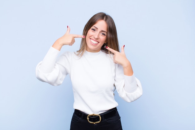 Young hispanic woman smiling confidently pointing to own broad smile, positive, relaxed, satisfied attitude on blue wall