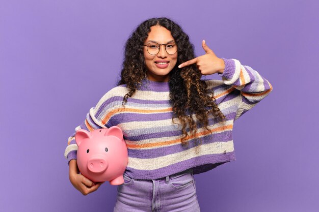 Young hispanic woman smiling confidently pointing to own broad smile, positive, relaxed, satisfied attitude. piggy bank concept