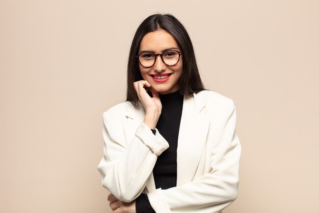 Young hispanic woman smiling, enjoying life, feeling happy, friendly, satisfied and carefree with hand on chin