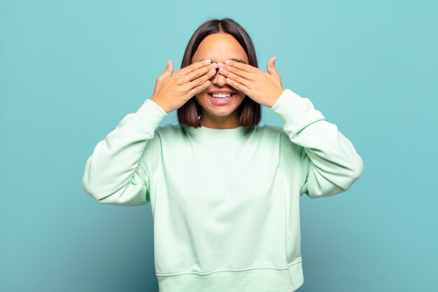 Young hispanic woman smiling and feeling happy, covering eyes with both hands and waiting for unbelievable surprise