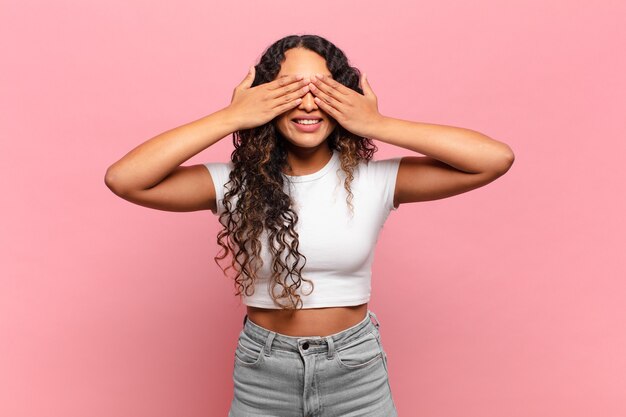 Young hispanic woman smiling and feeling happy, covering eyes with both hands and waiting for unbelievable surprise