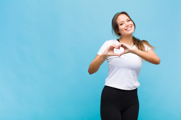 Young hispanic woman smiling and feeling happy, cute, romantic and in love, making heart shape with both hands