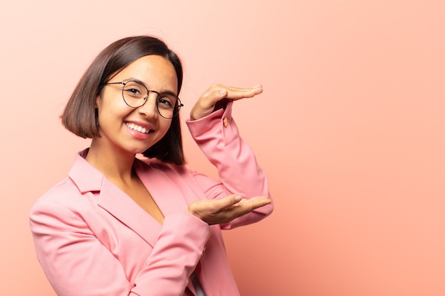 young hispanic woman smiling, feeling happy, positive and satisfied, holding or showing object or concept on copy space