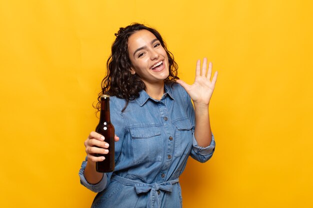 Young hispanic woman smiling happily and cheerfully, waving hand, welcoming and greeting you, or saying goodbye