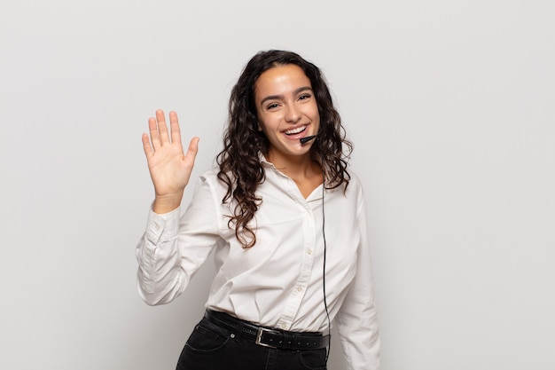 young hispanic woman smiling happily and cheerfully, waving hand, welcoming and greeting you, or saying goodbye