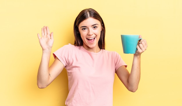 young hispanic woman smiling happily, waving hand, welcoming and greeting you. coffee cup concept