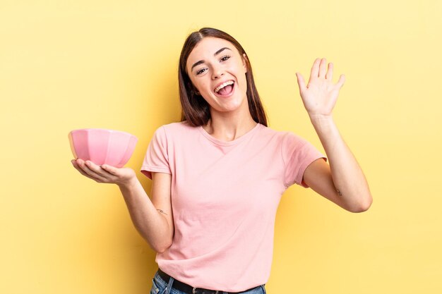 Young hispanic woman smiling happily waving hand welcoming and greeting you empty bowl concept