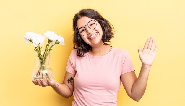 Giovane donna ispanica che sorride felicemente, agitando la mano, accogliendoti e salutandoti. concetto di vaso di fiori