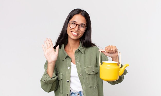 Young hispanic woman smiling happily, waving hand, welcoming and greeting you and holding a tea pot