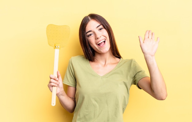 Giovane donna ispanica sorridente felicemente agitando la mano dandoti il benvenuto e salutandoti uccidi il concetto di mosche