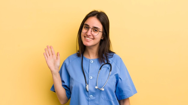 Foto giovane donna ispanica sorridente felicemente agitando la mano dandoti il benvenuto e salutandoti concetto di infermiera