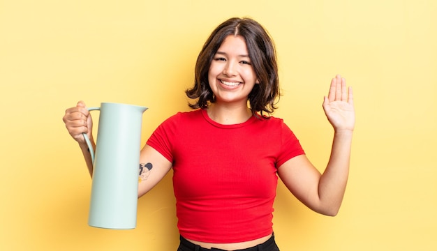 Молодая латиноамериканская женщина счастливо улыбается, машет рукой, приветствует и приветствует вас. концепция термоса