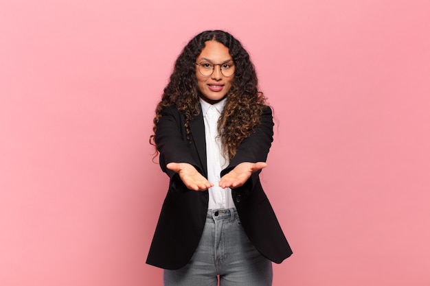 Young hispanic woman smiling happily with friendly, confident, positive look, offering and showing an object or concept. business concept