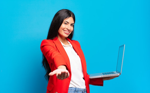 Young hispanic woman smiling happily with friendly, confident, positive look, offering and showing an object or concept. laptop concept