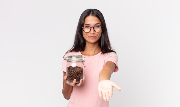 Giovane donna ispanica che sorride felicemente con amichevole e offrendo e mostrando un concetto e tenendo in mano una bottiglia di chicchi di caffè