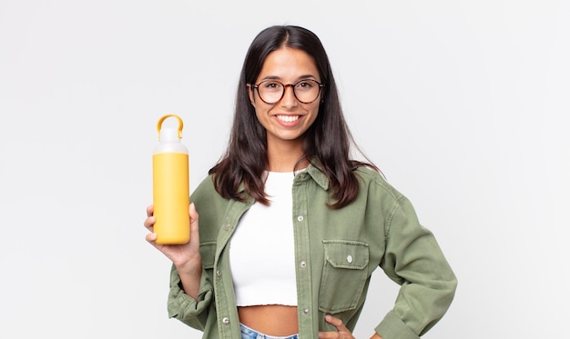 Young hispanic woman smiling happily with a hand on hip and confident and holding a coffee thermos