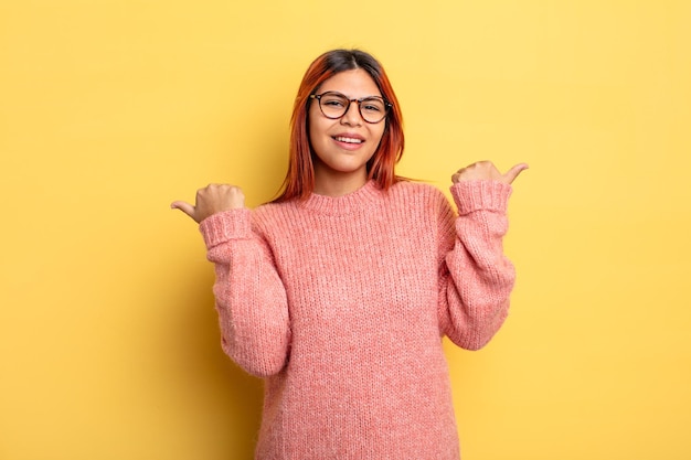 嬉しそうに笑って幸せそうに見える若いヒスパニック系の女性は、両方の親指を上に向けてのんきで前向きに感じています