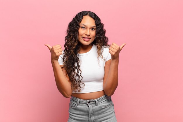 young hispanic woman smiling joyfully and looking happy, feeling carefree and positive with both thumbs up