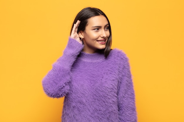 Young hispanic woman smiling, looking curiously to the side, trying to listen to gossip or overhearing a secret