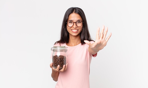 Young hispanic woman smiling and looking friendly, showing number five and holding a coffee beans bottle