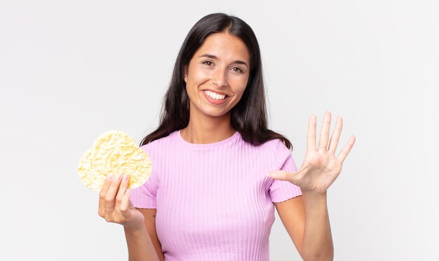 Young hispanic woman smiling and looking friendly, showing number five and holding a rice cookie. diet concept