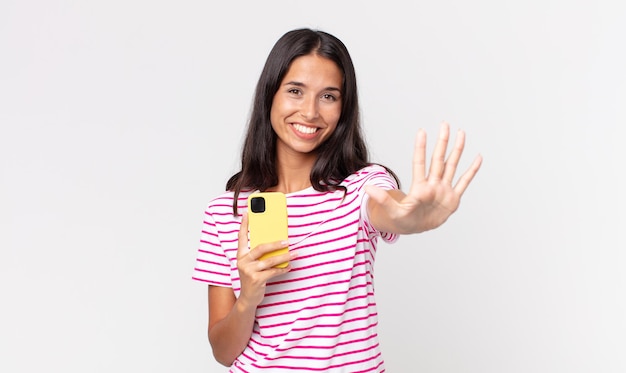 Young hispanic woman smiling and looking friendly, showing number five and holding a smartphone