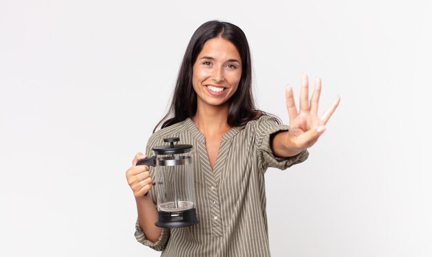 Молодая латиноамериканская женщина улыбается и выглядит дружелюбно, показывает номер четыре и держит ручную кофеварку