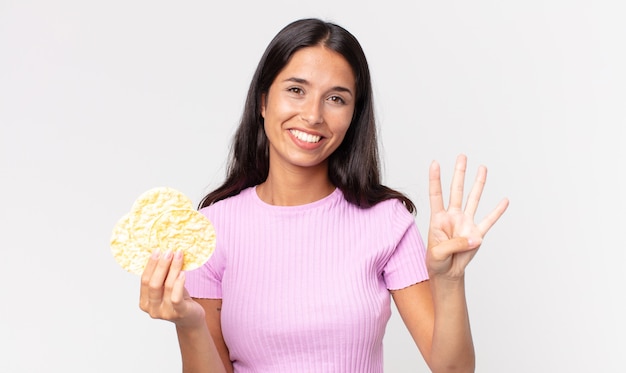 Giovane donna ispanica sorridente e dall'aspetto amichevole, mostrando il numero quattro e tenendo in mano un biscotto di riso. concetto di dieta