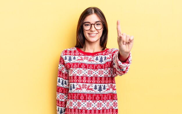 Young hispanic woman smiling and looking friendly, showing number one. chistmas and new year concept