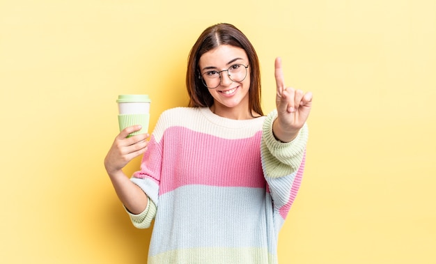 Young hispanic woman smiling and looking friendly, showing number one. take away coffee concept