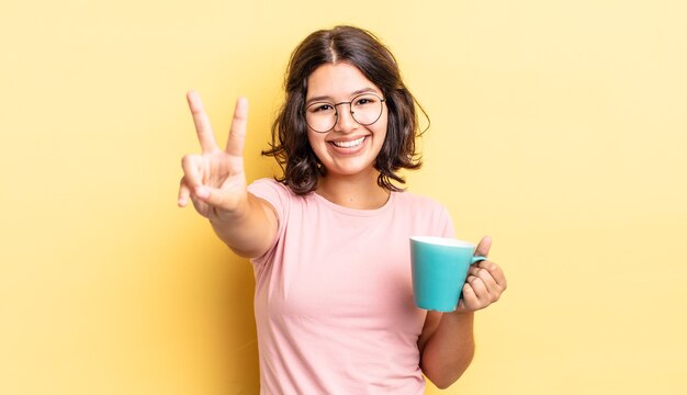 Young hispanic woman smiling and looking friendly, showing number two. coffee mug concept