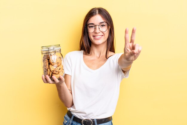 Young hispanic woman smiling and looking friendly, showing number two. cookies bottle concept