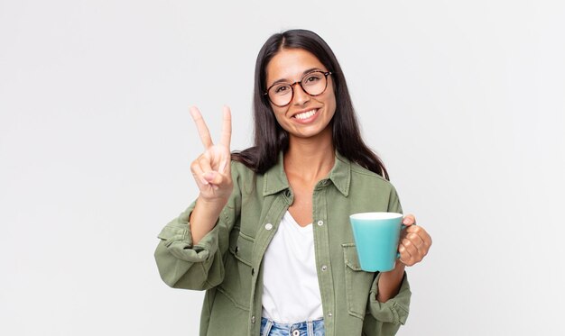 Young hispanic woman smiling and looking friendly, showing number two and holding a coffee mug