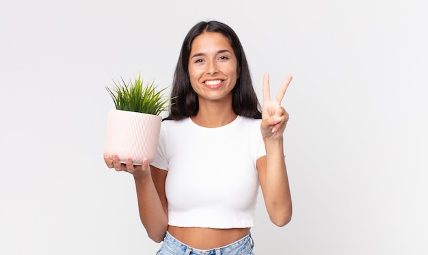 young hispanic woman smiling and looking friendly, showing number two and holding a decorative house plant