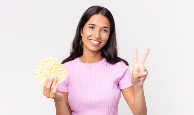 Young hispanic woman smiling and looking friendly, showing number two and holding a rice cookie. diet concept