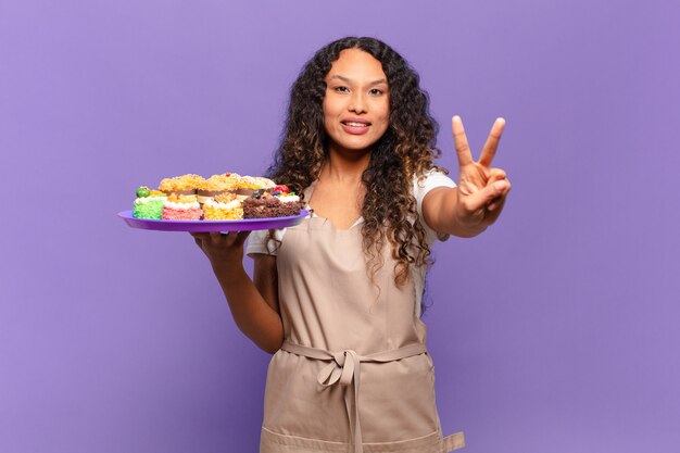 Young hispanic woman smiling and looking friendly, showing number two or second with hand forward, counting down. cooking cakes concept