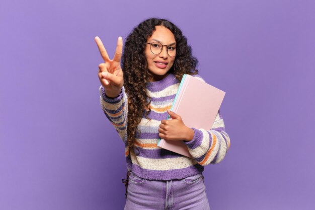 Young hispanic woman smiling and looking happy carefree and\
positive gesturing victory or peace with one hand student\
concept